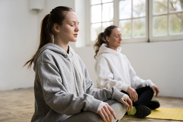 A teenager wearing a grey hoodie sits cross legged on a yoga mat, meditating